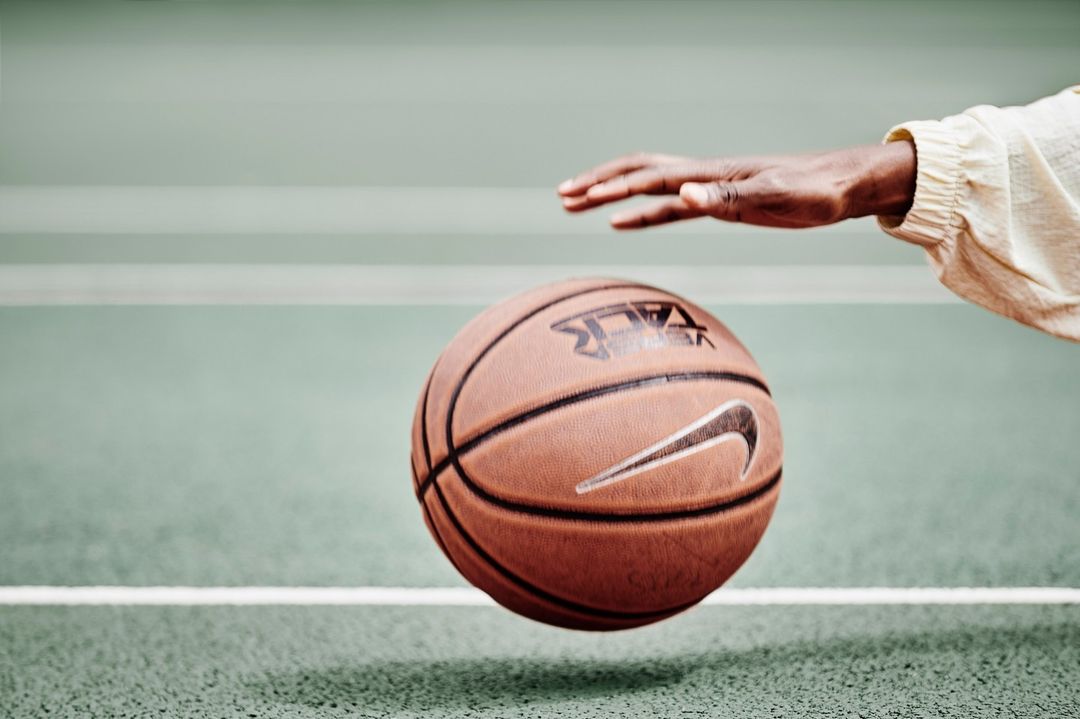 An arm reaches for an airborne basketball with a visible Nike logo, against a green and white court backdrop, showcasing sports action.