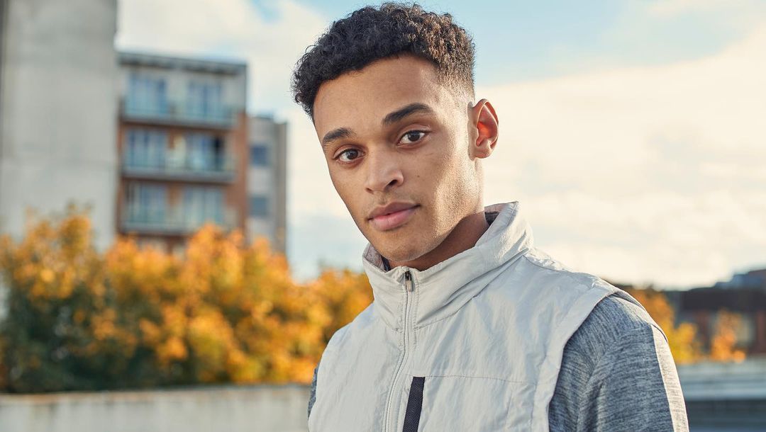 Portrait of a young adult male wearing a grey shirt and a vest outdoors, with a building and foliage in the background on a sunny day.