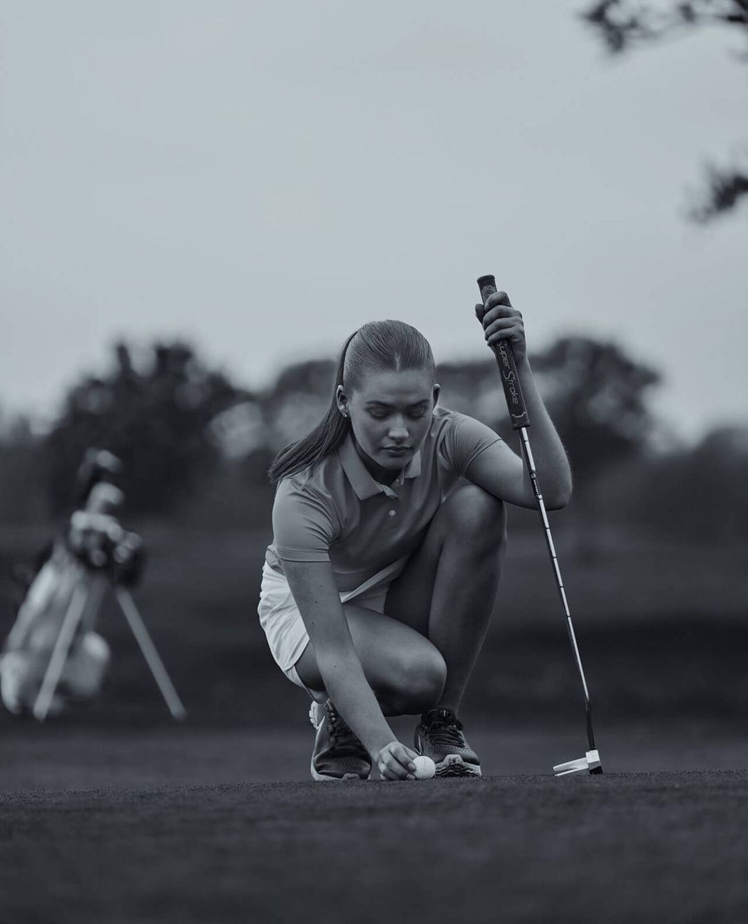 Young woman places golf ball on green course with golf club and bag in the background, set against an overcast sky.