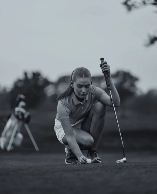 Young woman places golf ball on green course with golf club and bag in the background, set against an overcast sky.