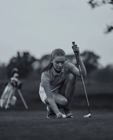 Young woman places golf ball on green course with golf club and bag in the background, set against an overcast sky.