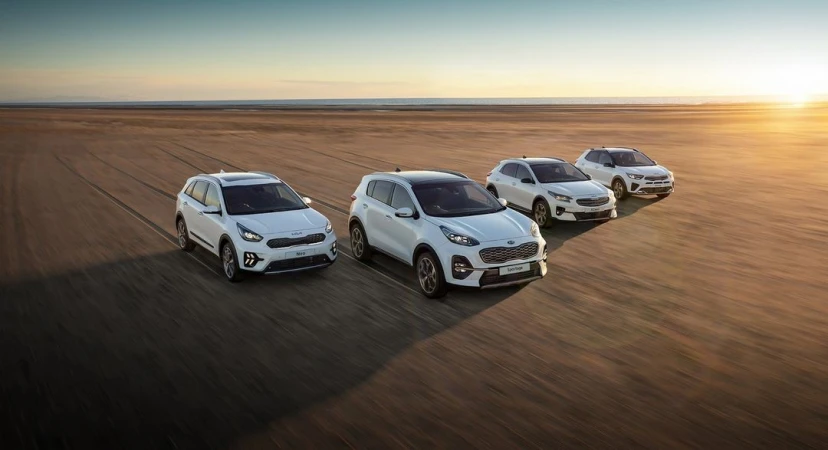 Four white Kia SUVs are parked on a tan beach at sunset. The scene is well-lit, highlighting the vehicle designs against the bright sky.