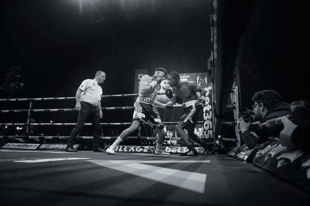 Monochrome shot of two adult male boxers in boxing ring in combat, with referee and photographer, bathed in bright arena lighting.