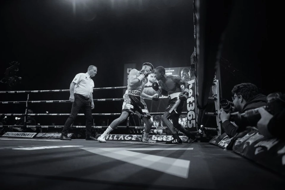 Monochrome shot of two adult male boxers in boxing ring in combat, with referee and photographer, bathed in bright arena lighting.