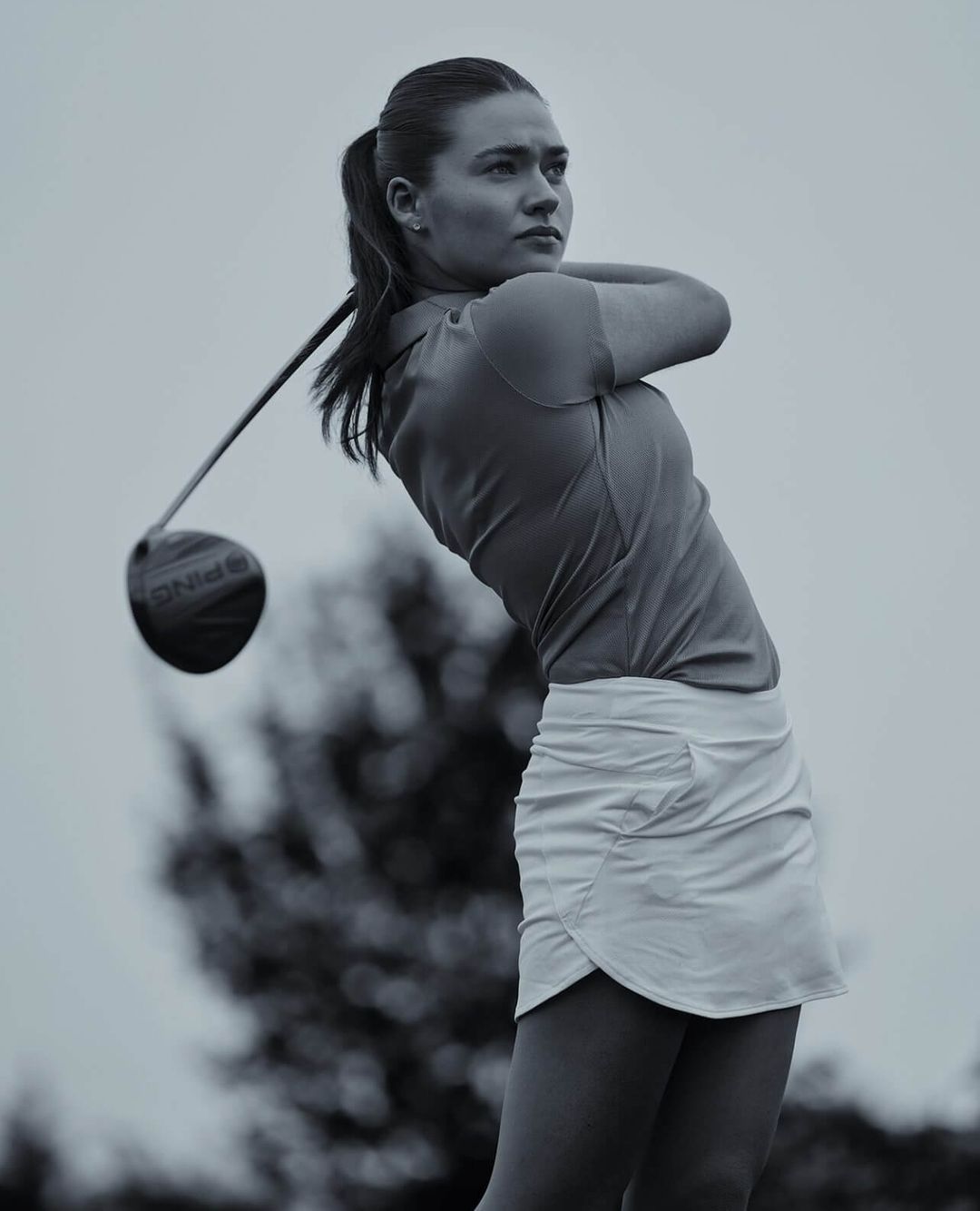 A woman in a sporty golf outfit is captured in motion, swinging a golf club on a grassy field, set against a bright, overcast sky.