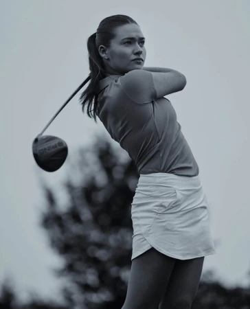 A woman in a sporty golf outfit is captured in motion, swinging a golf club on a grassy field, set against a bright, overcast sky.