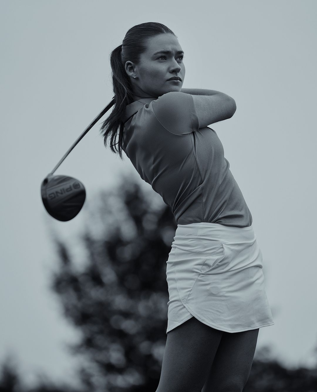 A woman golfer in a golf swing on a golf course, wearing a white skirt and orange shirt, swinging a Ping driver with trees behind her.
