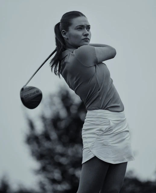A woman golfer in a golf swing on a golf course, wearing a white skirt and orange shirt, swinging a Ping driver with trees behind her.