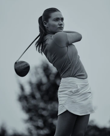 A woman golfer in a golf swing on a golf course, wearing a white skirt and orange shirt, swinging a Ping driver with trees behind her.