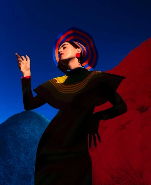 Fashionable woman in blue-and-red striped hat and angular dress poses against vibrant backdrop of red and blue contrasting color fields.