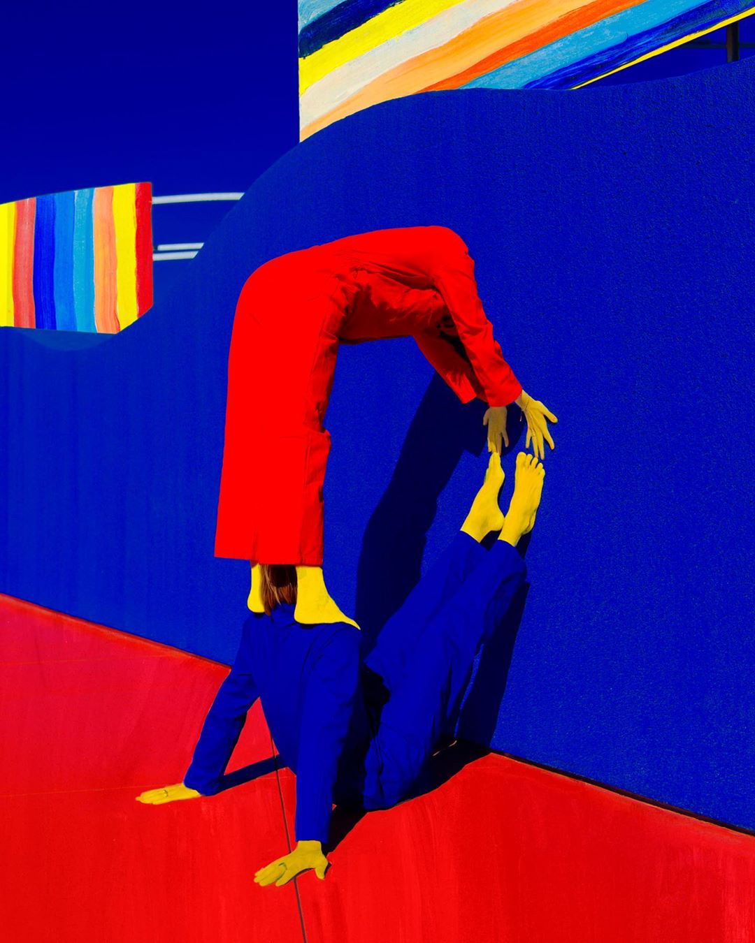Abstract composition with two people in full-body suits creating an acrobatic arch against a blue backdrop and a red floor. Bright, saturated colors.