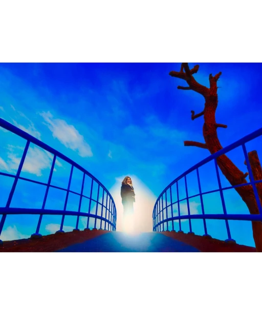 Woman standing on a blue bridge against a vibrant blue sky with light clouds, with a red-brown tree on the side, creating a surreal scene.