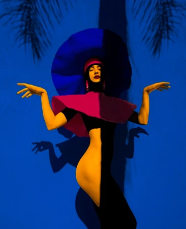 Stylized fashion portrait of a woman in a two-tone body suit, wide-brimmed hat, and palm tree shadows against a blue background.