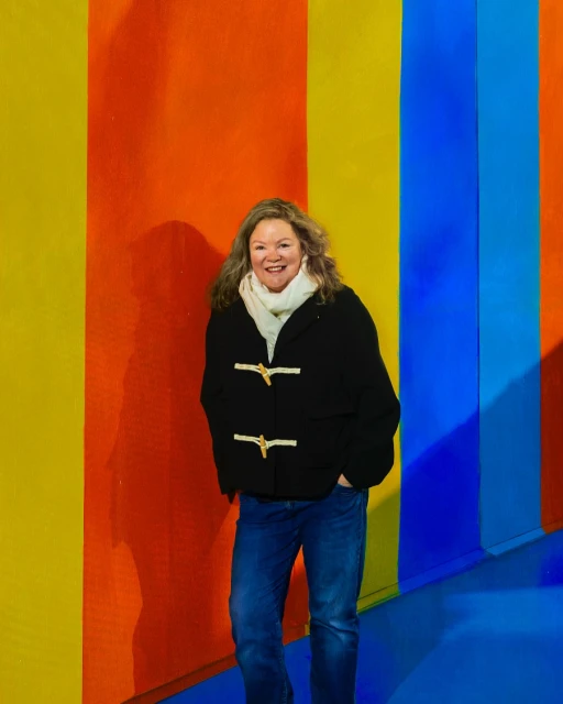 Adult woman with long brown hair stands in front of a striped wall, wearing a duffle coat, scarf and jeans. Casual portrait.
