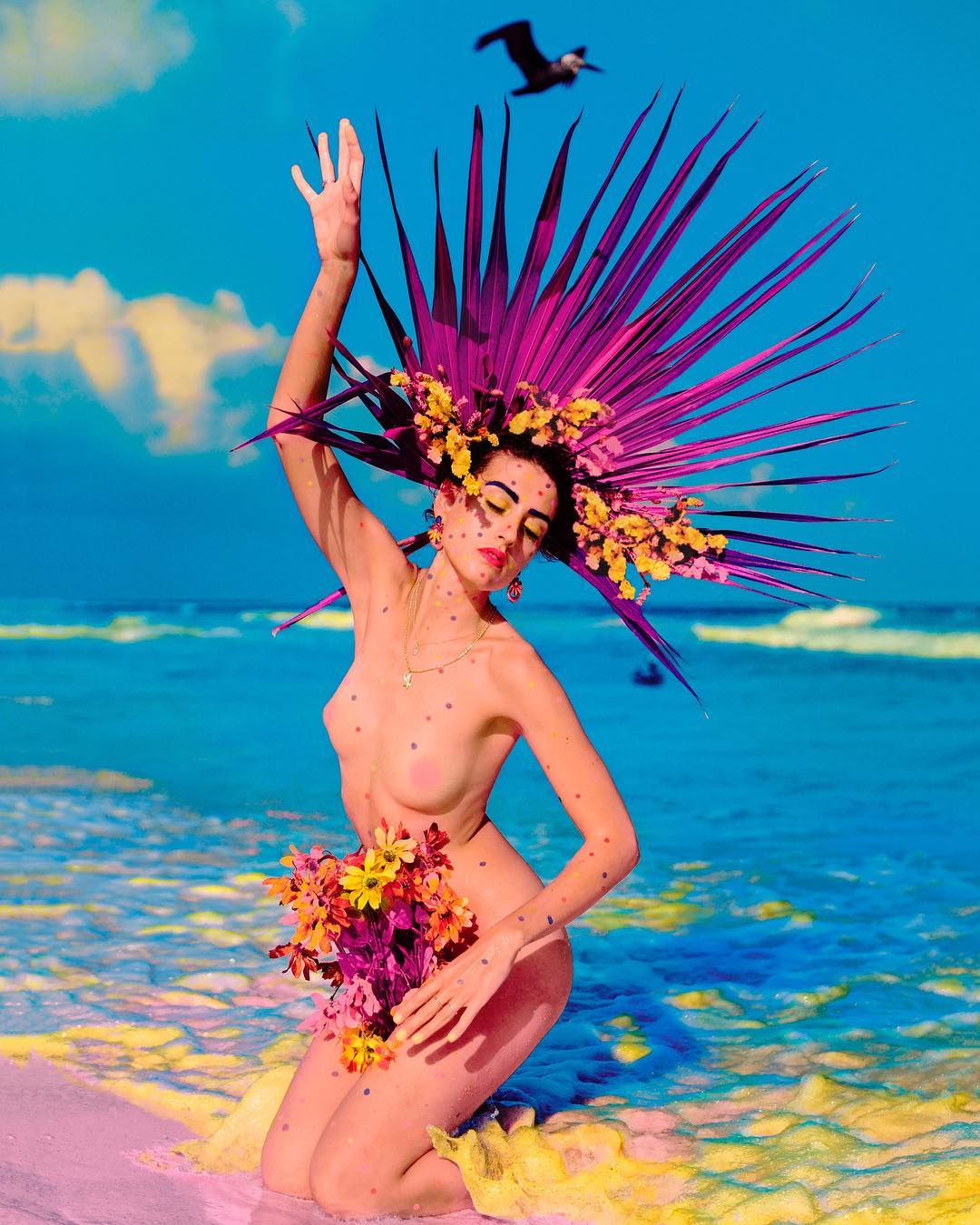 Woman kneeling on a beach with a floral headdress and flower bouquet, with a bird flying overhead, against a blue sky and ocean backdrop.