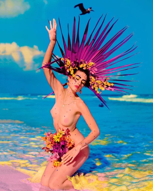 Woman kneeling on a beach with a floral headdress and flower bouquet, with a bird flying overhead, against a blue sky and ocean backdrop.