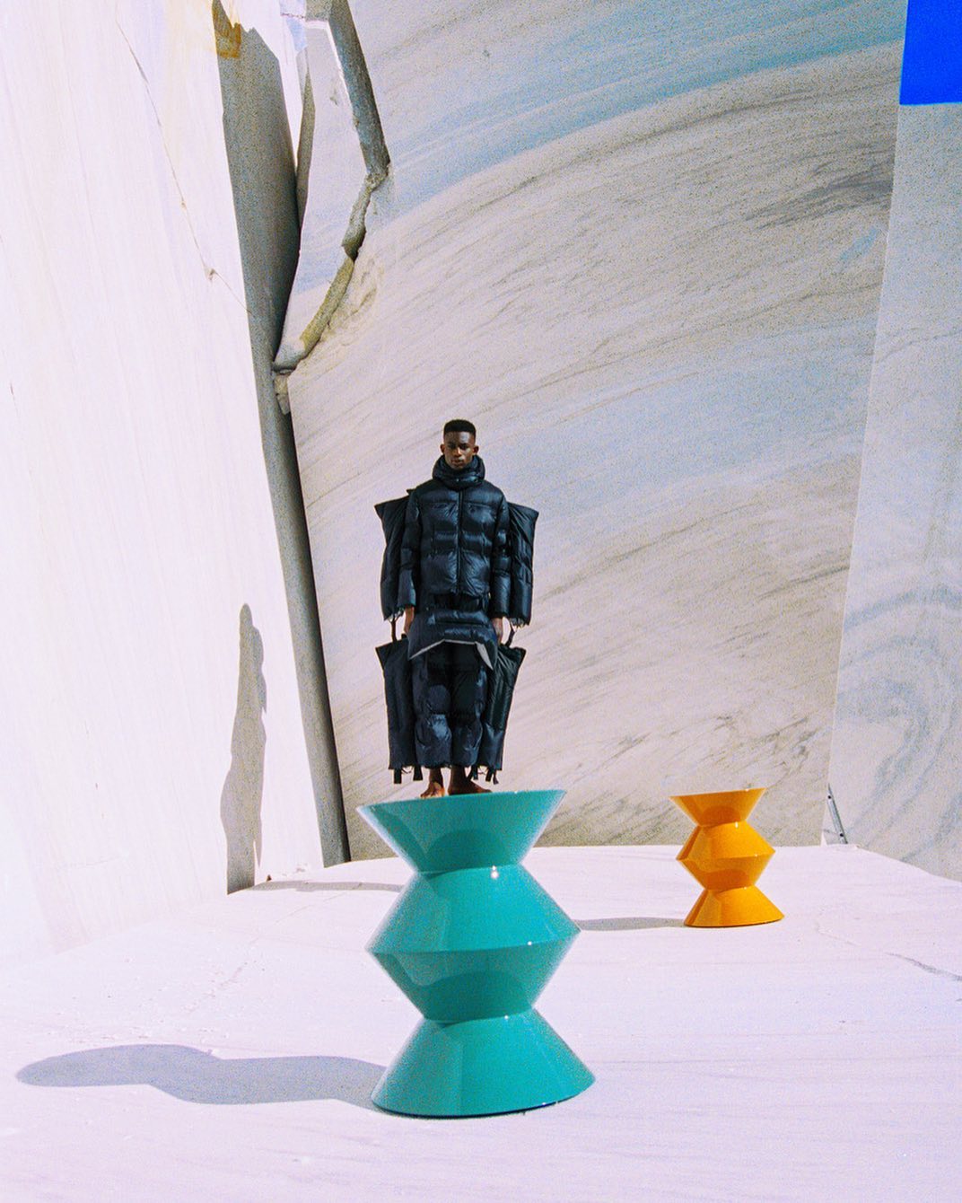 A person in a black puffer suit stands on a teal hourglass stool with a yellow hourglass stool behind them, set against a marble background.