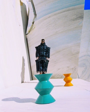 A person in a black puffer suit stands on a teal hourglass stool with a yellow hourglass stool behind them, set against a marble background.