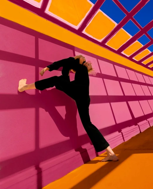A woman in a black jumpsuit performs a contortionist pose against a pink wall with an orange floor and blue ceiling.