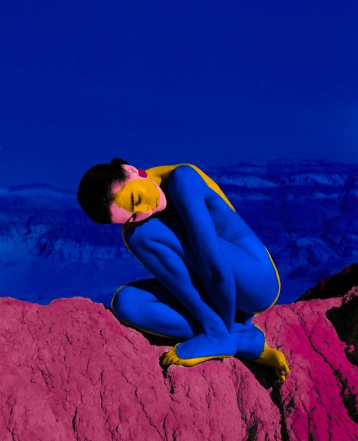 Woman with blue body paint in a crouching pose on a reddish-pink textured surface with a dark blue and purple background.