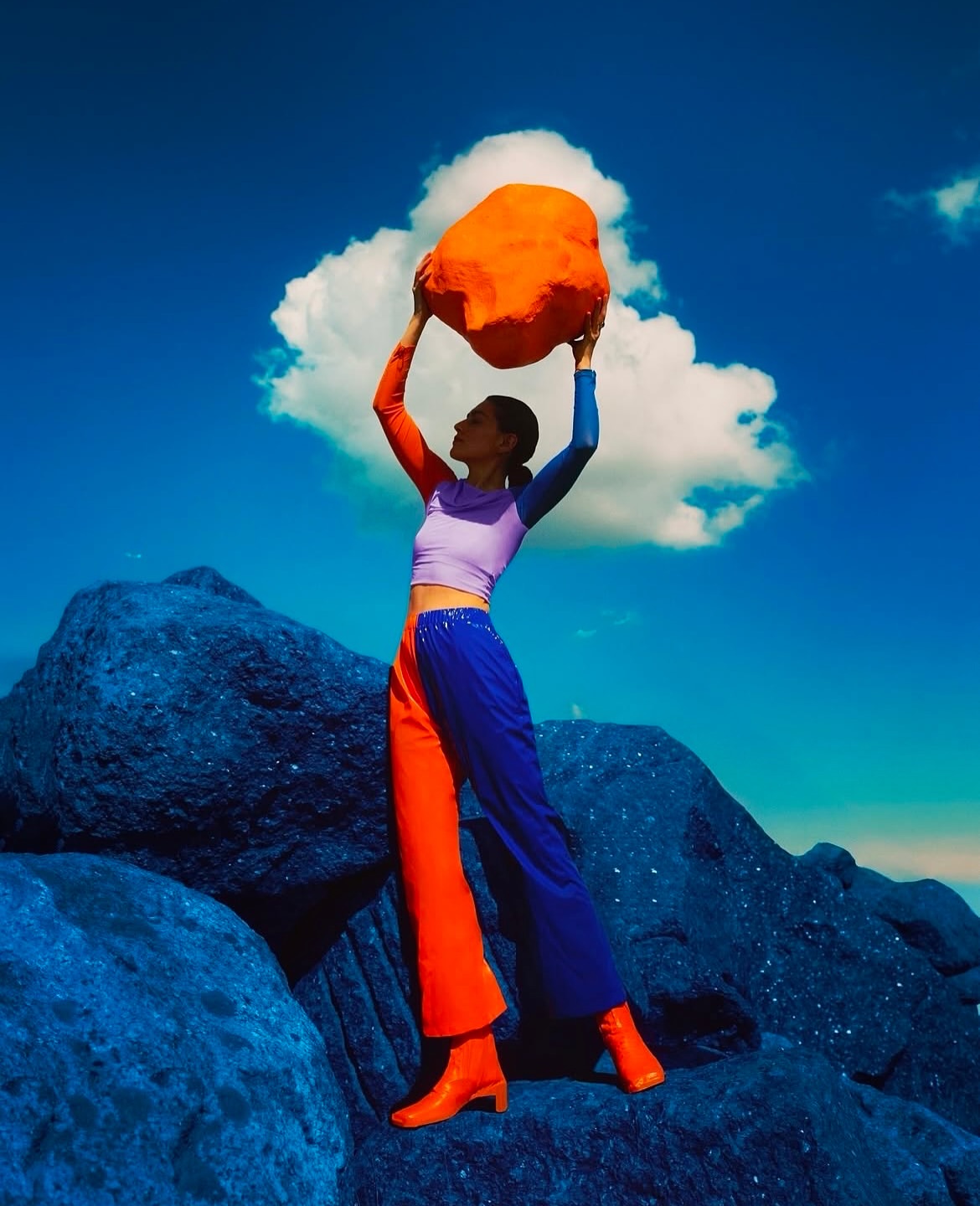 Woman in colour-blocked clothing stands among blue rocks holding an orange, abstract rock under a bright blue sky with a white cloud.