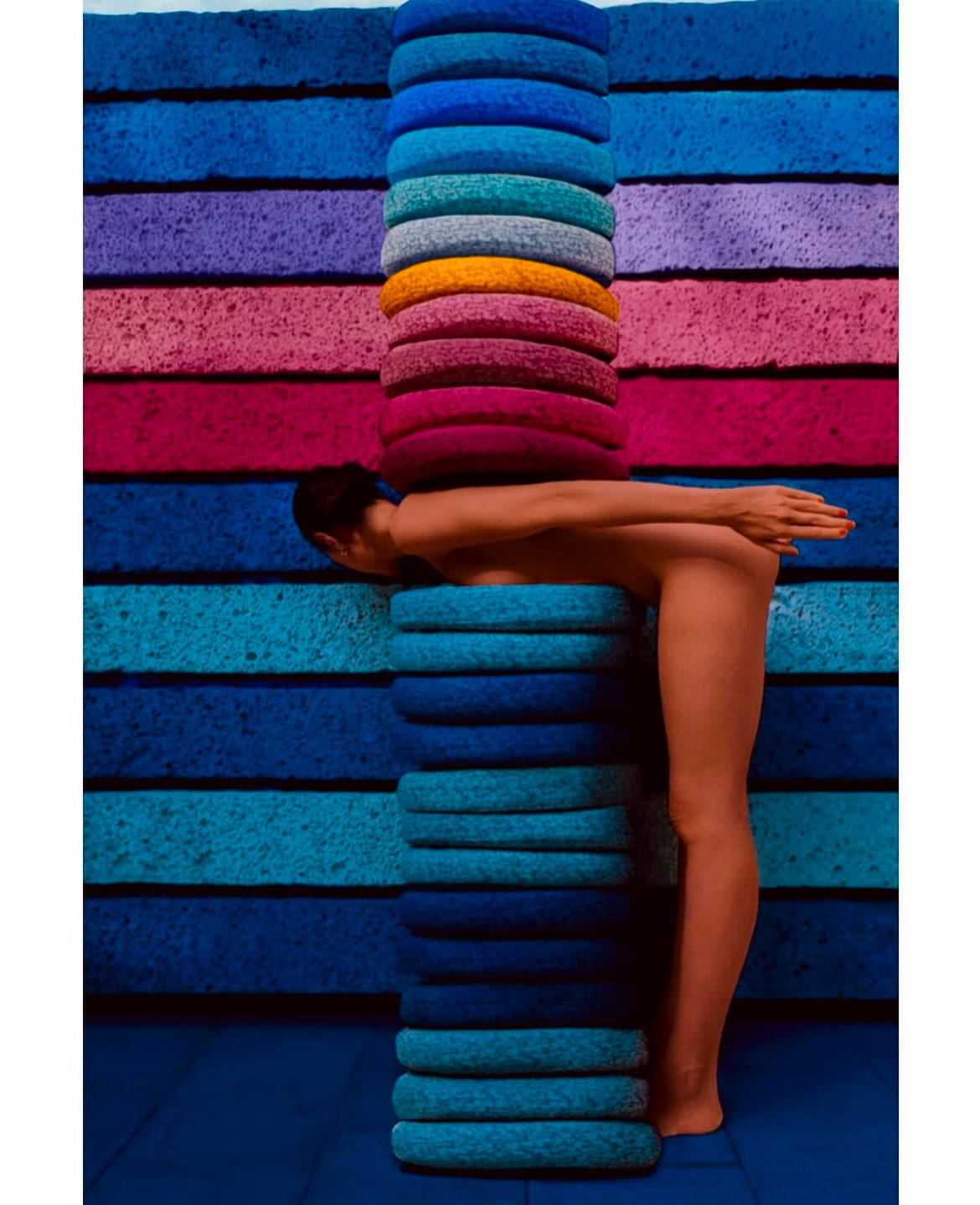 Artistic nude portrait of an adult woman posing between colorful stacked mats on a blue floor in a studio with creative lighting.