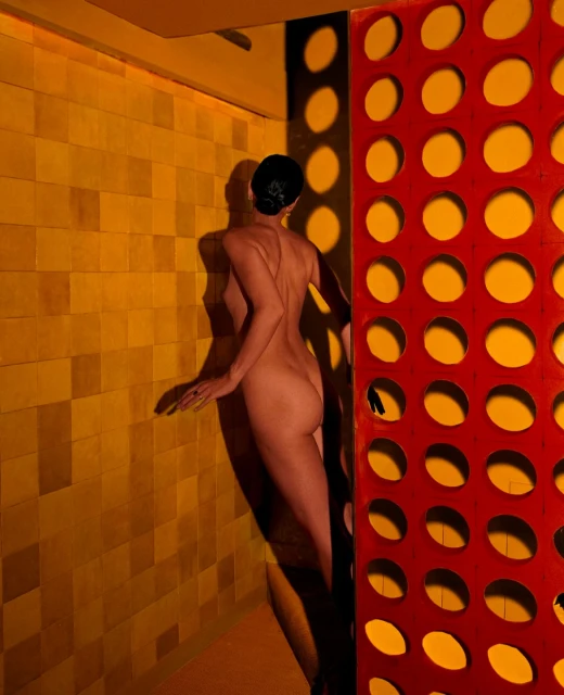 An adult female stands next to a decorative wall with circular openings in a warm-toned indoor setting, casting a shadow on a tiled wall.
