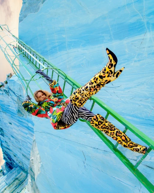 Fashion shot of a woman on a ladder wearing animal print boots, zebra pants, and a floral top against a light blue backdrop.