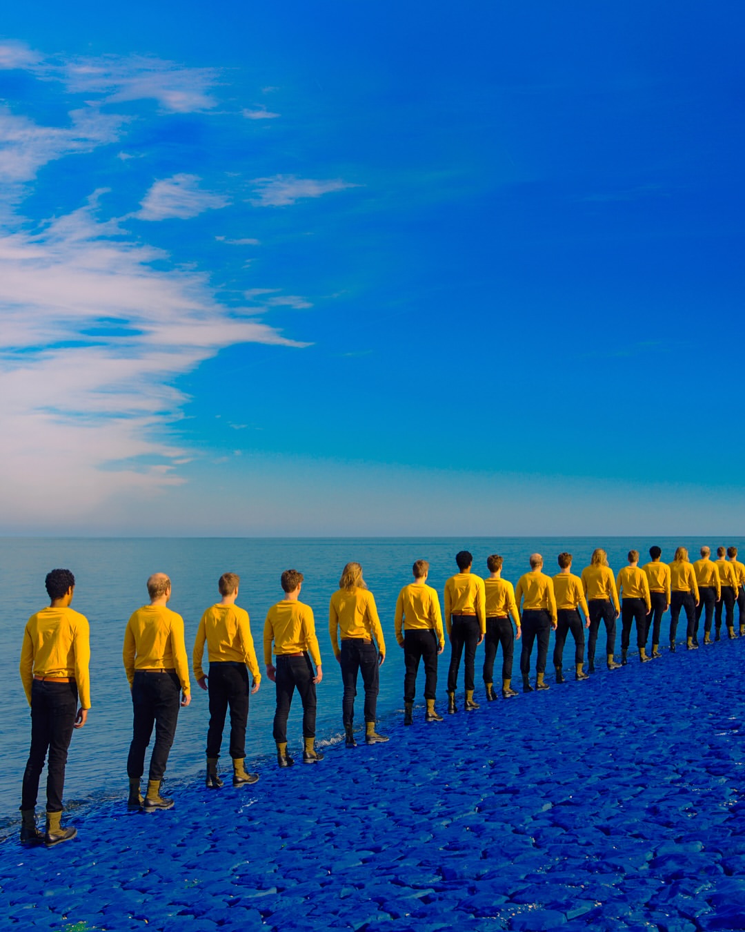 A long row of people standing at the water's edge, all wearing yellow shirts and black pants, creates a striking visual against the blue sea and sky.