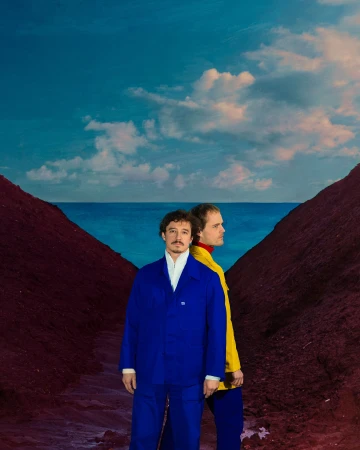 Two men stand in striking attire against an otherworldly backdrop of a blue sky, distant water, and red earth formations, creating a surreal scene.
