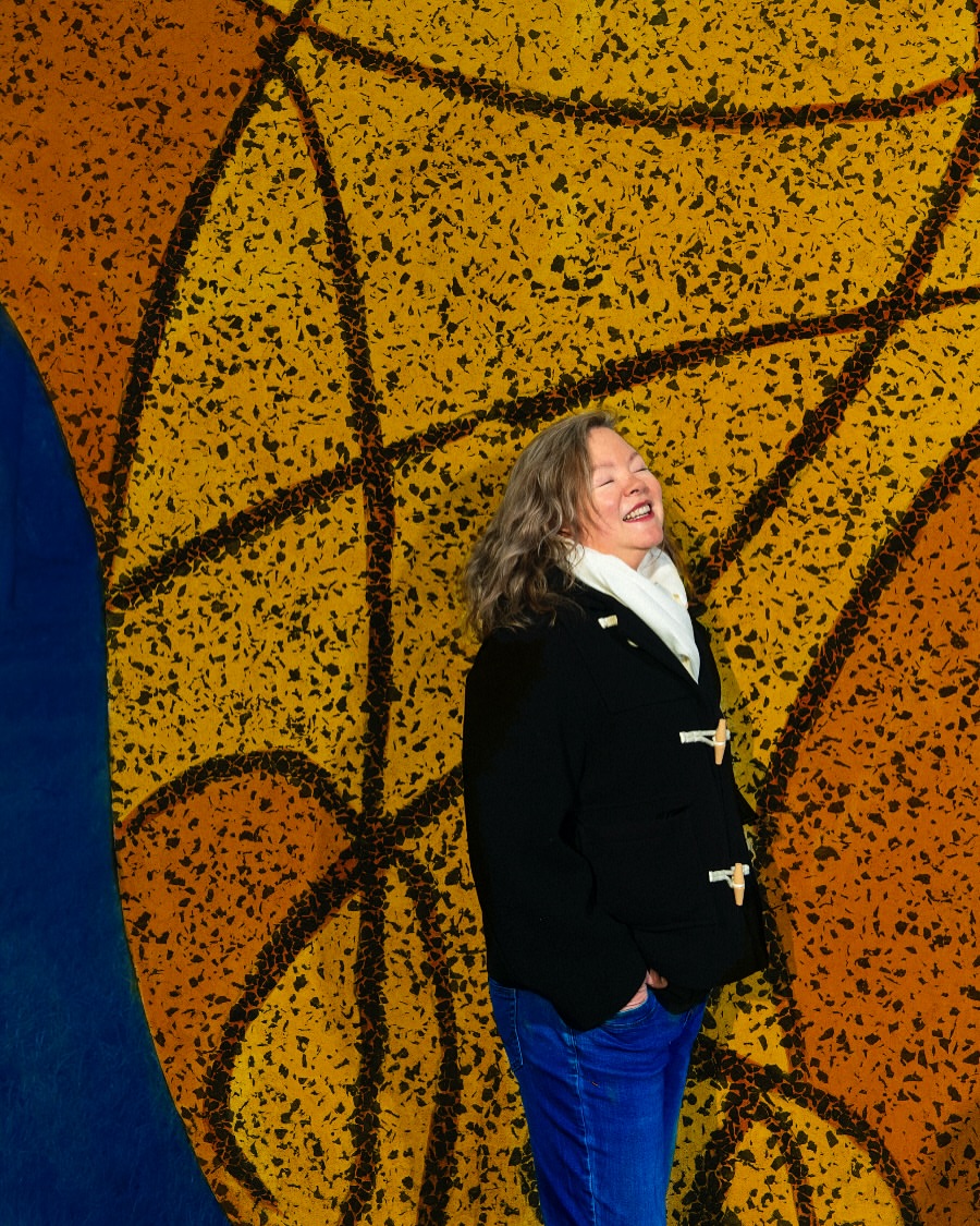 Portrait of smiling mature woman in black duffel coat, blue jeans, and white scarf, standing in front of yellow abstract art.