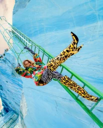 Model in vibrant floral jacket and leopard print boots climbing a green ladder against a bright blue marble quarry wall.