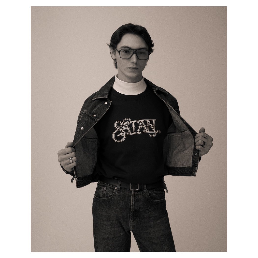 Black and white portrait of a stylish young adult male wearing a black t-shirt with 'SATAN' printed on it, denim jacket, and glasses.