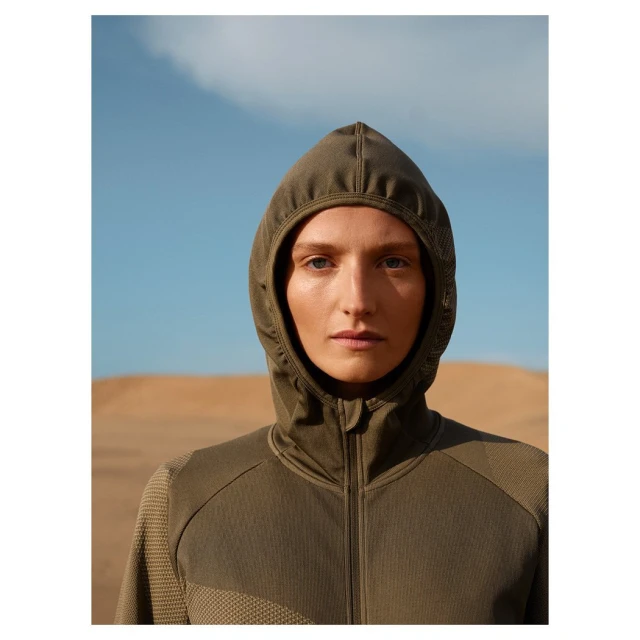 Portrait of an adult female wearing a green hooded jacket, set against a backdrop of a sand dune and a clear blue sky.