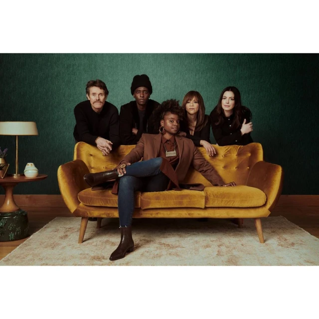 Five adults on a yellow couch against a green wallpapered wall. One woman sits, while two women and two men stand behind the couch.