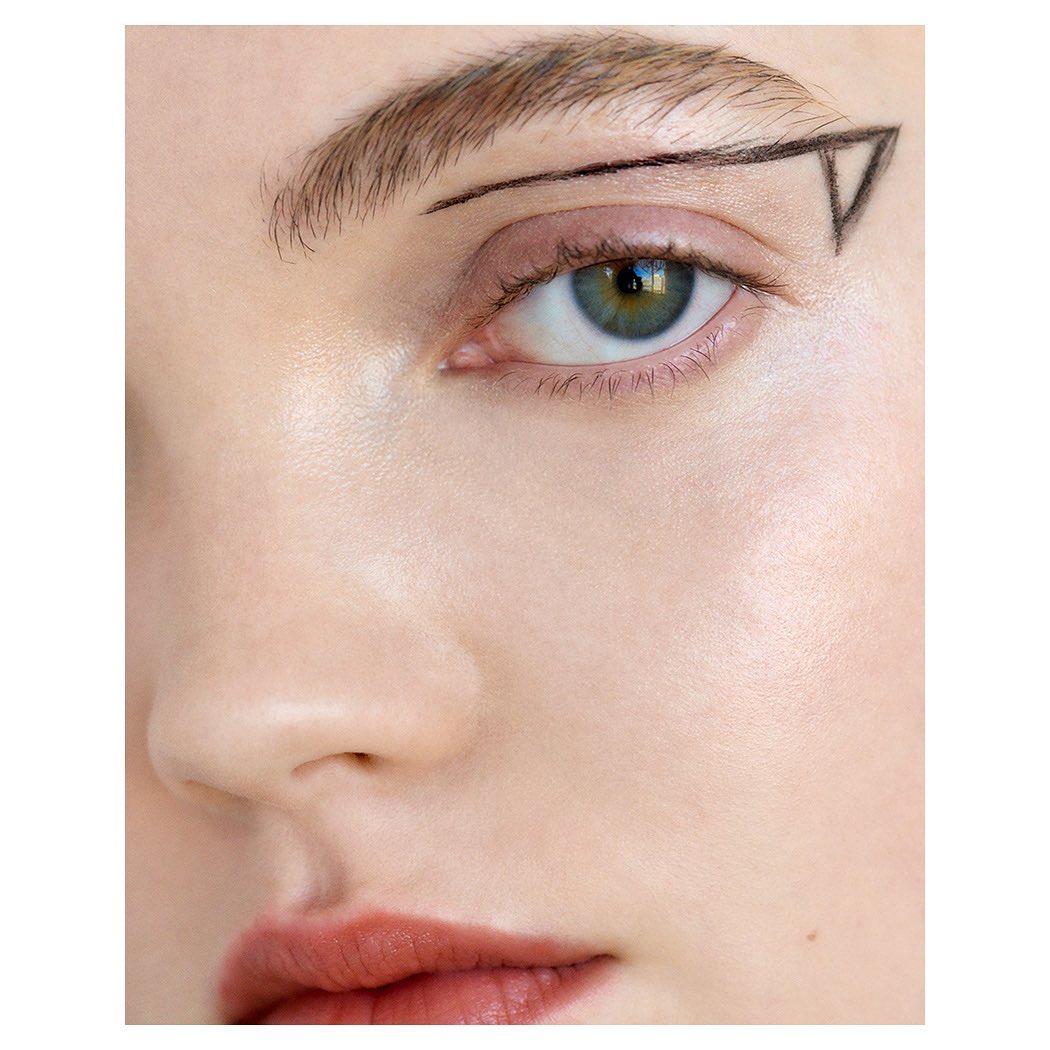 Close-up of a fair-skinned adult female face with makeup and a black line art graphic near her left eyebrow in a square format.