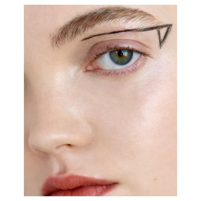 Close-up of a fair-skinned adult female face with makeup and a black line art graphic near her left eyebrow in a square format.