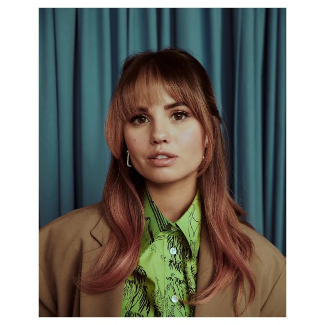 Portrait of Debby Ryan wearing a brown blazer and patterned green blouse in front of teal curtains in a close-up studio shot.