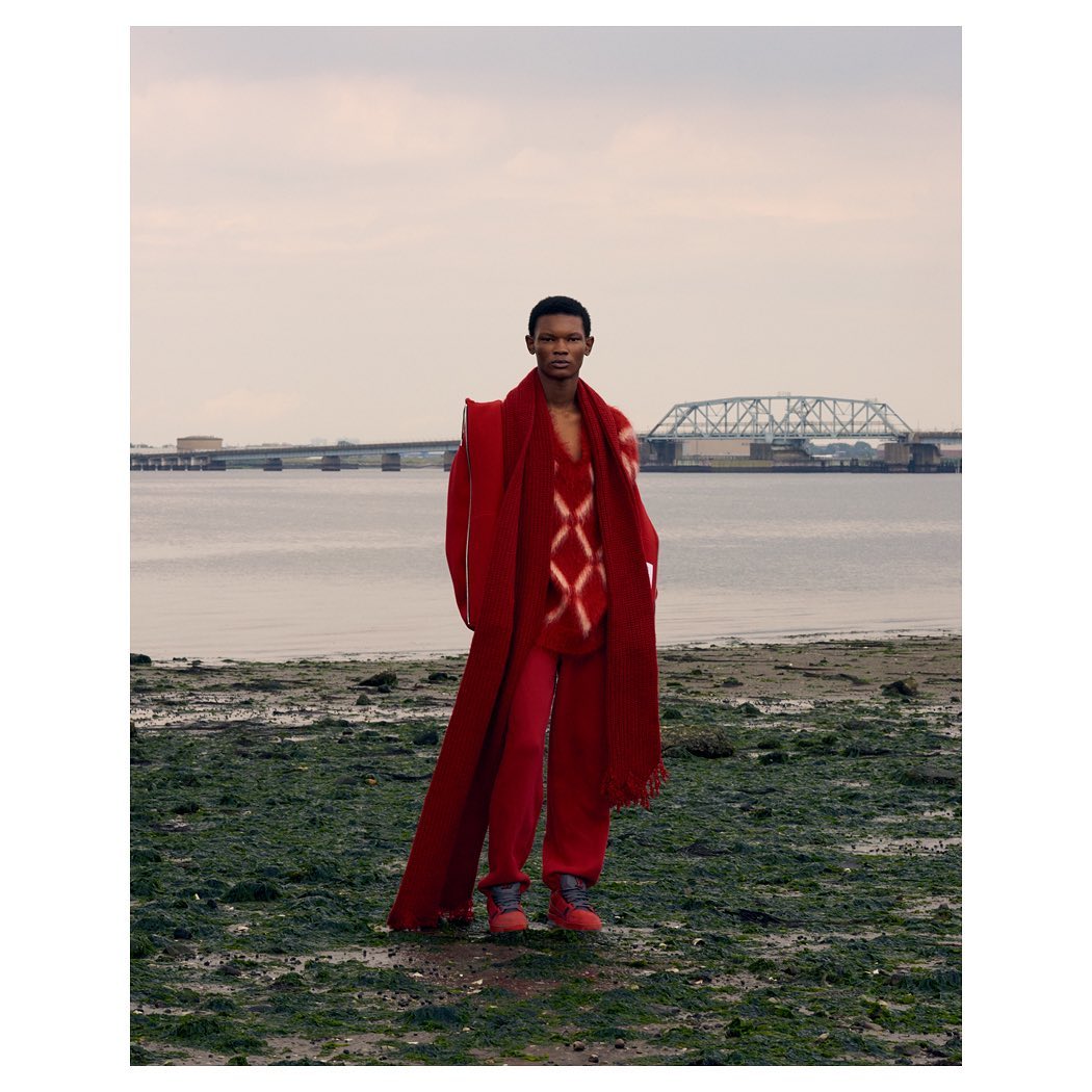 Fashion shot of a Black male model in red coat, sweater, and pants posing on a beach with a bridge and water in the background.