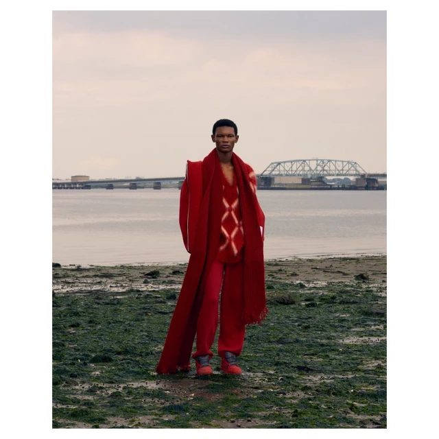 Fashion shot of a Black male model in red coat, sweater, and pants posing on a beach with a bridge and water in the background.