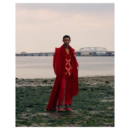 Fashion shot of a Black male model in red coat, sweater, and pants posing on a beach with a bridge and water in the background.
