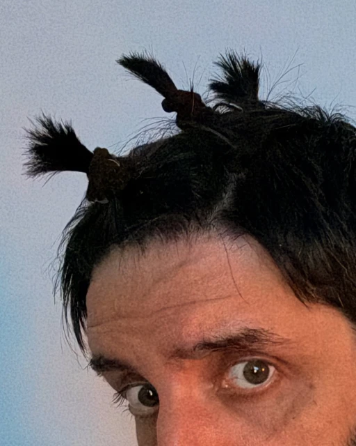 Close-up of a man with dark hair styled into two whimsical top knots resembling animal ears, set against a soft, light blue background.