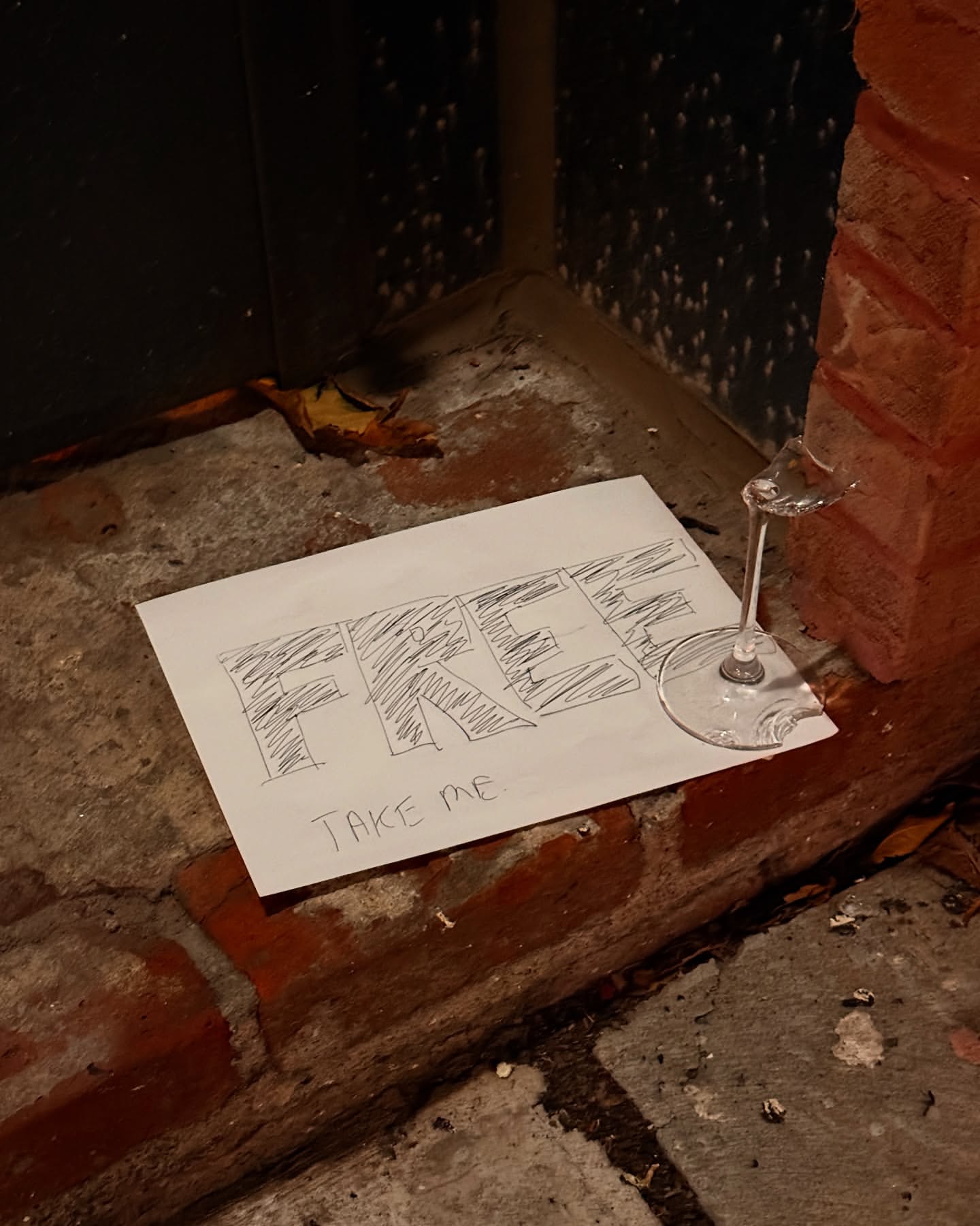Handwritten 'Free Take Me' sign on brick floor with a glass on the edge of the paper, next to a dark blue surface.