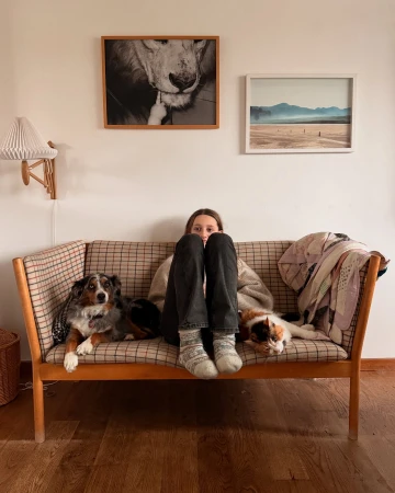 A young woman relaxes on a plaid couch with her legs up, accompanied by an Australian Shepherd dog and a calico cat in a cozy home setting.