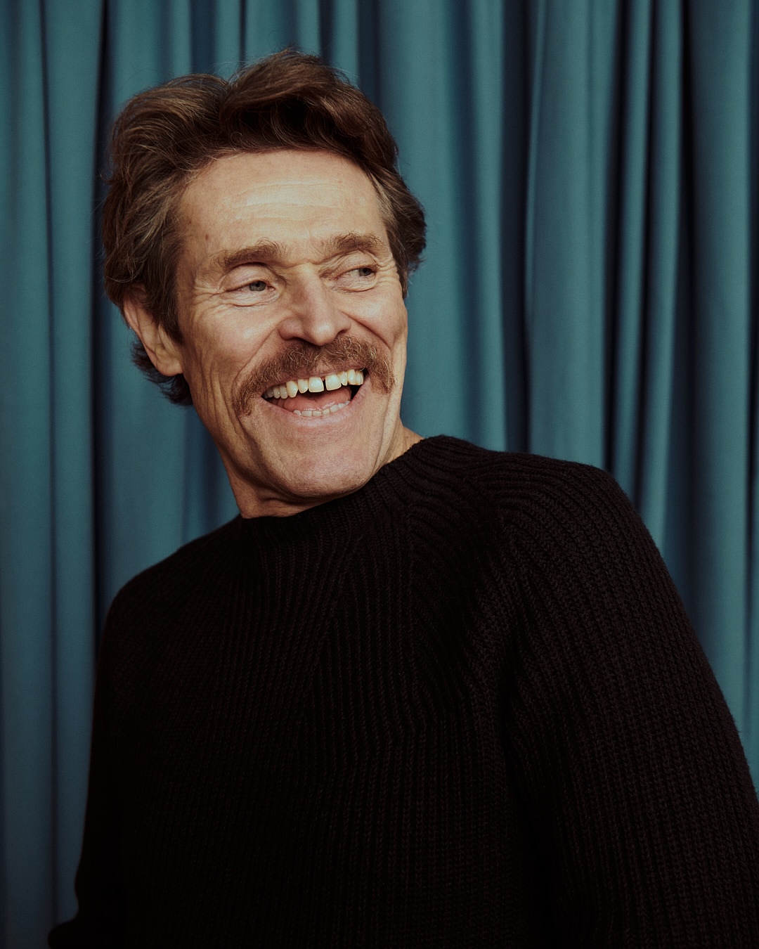 Portrait of Willem Dafoe, an adult man with brown hair, mustache, and a black sweater, smiling against a teal curtain backdrop.