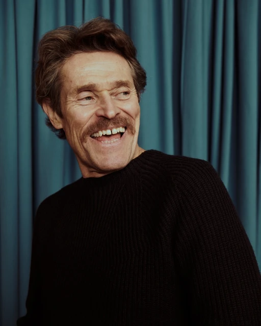 Portrait of Willem Dafoe, an adult man with brown hair, mustache, and a black sweater, smiling against a teal curtain backdrop.