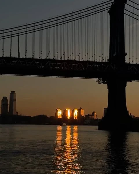 Silhouette of the Manhattan Bridge at sunset with golden light reflecting off the buildings and water.