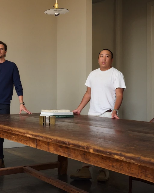Two adult men stand behind a long wooden table in a bright interior space, with books and brass objects arranged on the table surface.