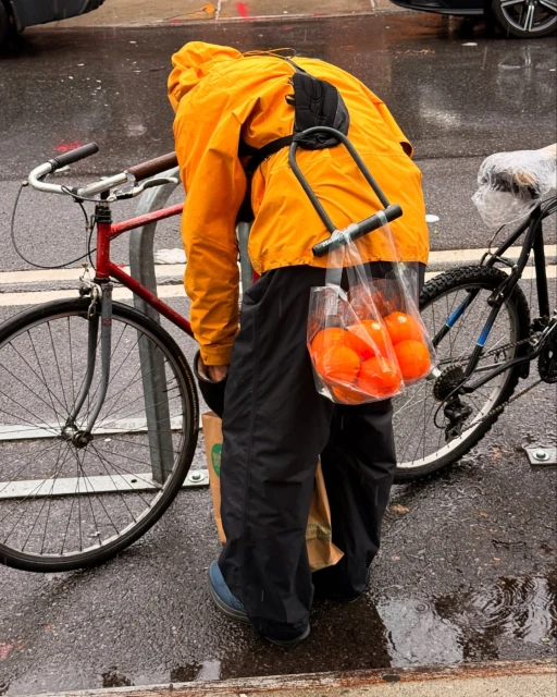 Person in yellow raincoat transports oranges on bike rack through wet urban environment. Second bicycle and shopping bag visible.