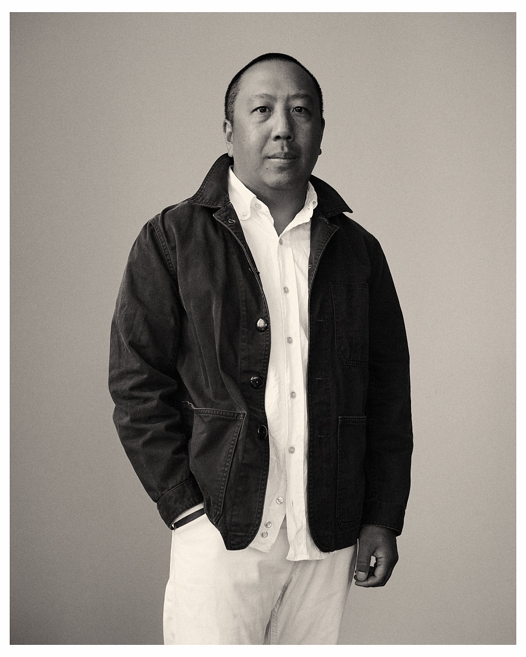 Portrait of an adult man with short dark hair, wearing a white shirt, dark jacket, and light-colored trousers, against a neutral background.